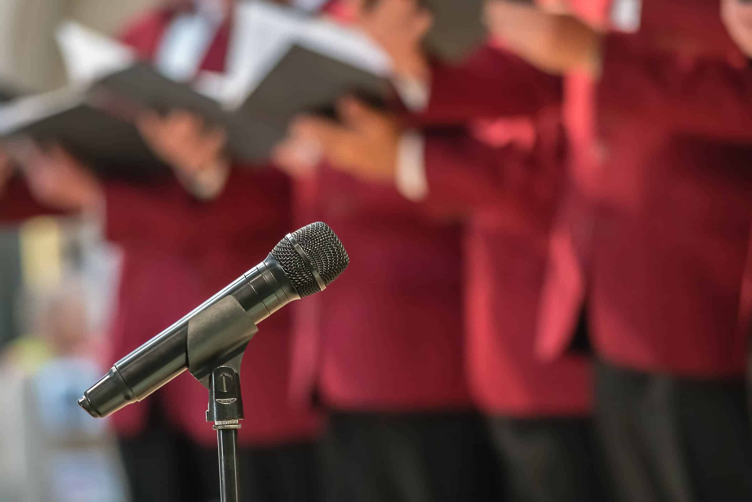 Decorative header photo of a microphone with men in red jackets out of focus in the back ground.